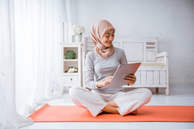 Müslüman tesettürlü kadın egzersiz evinde antrenman yapıyor. Odadaki döşeklerin üzerindeki tabletten mutlu bir şekilde ders veriyor.