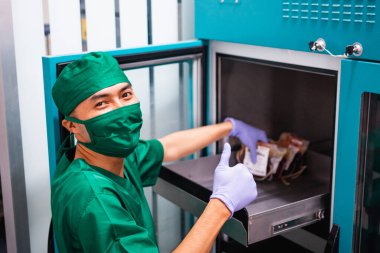 Erkek hastane çalışanı üniformalı ve baş parmaklı bir maske takarken depoda kan torbası saklıyor.