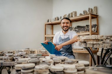 Erkek girişimci kameraya gülümsüyor. Taş el sanatları deposunda elinde bir tahta ve kalemle oturuyor.