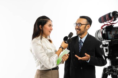 Gözlüklü yetişkin bir adam beyaz bluzlu kadın gazeteciyle siyah bir mikrofon kullanarak konuşuyor ve bir set kamera tarafından kaydediliyor.