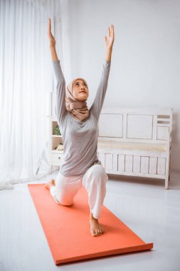 Asyalı Müslüman kadın eğitmen tesettür giyip Anjaneyasana yoga pozu veriyor turuncu pilates yatağında.