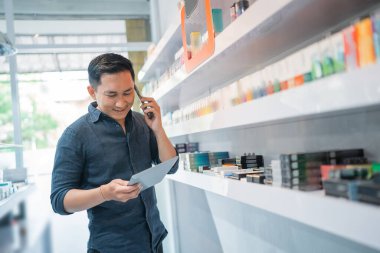 Erkek esnaf, telefon ederken ve dijital tablete bakarken mağazanın rafının yanında duruyor.