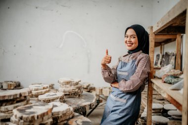 Asyalı tesettürlü Müslüman kadın, taş el sanatları atölyesinde rafa yaslanmış baş parmaklarıyla gülümsüyor.