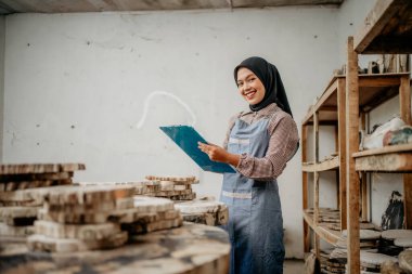 Asyalı peçeli kadın kameraya gülümsüyor. Elinde taş sanatları atölyesinde bir pano var.