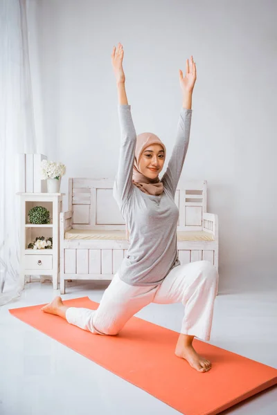 亚洲穆斯林女教师穿着头巾T恤衫辅导Anjaneyasana瑜伽在橙色普拉提床垫上 微笑着看着房间里的相机 — 图库照片
