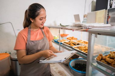 Garson, geleneksel bir yemek tezgahının panosunda kağıt kontrol listesi kullanarak hazır bir menüye bakıyor.