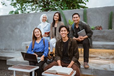 Bir grup gülümseyen genç öğrenci, üniversitede kitap ve dizüstü bilgisayarla birlikte mutlu bir şekilde bir araya geldiler.