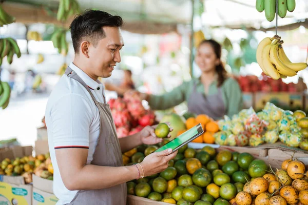 男の果物屋はオレンジをチェックしながらデジタルタブレットを見ている女性の売り手は背景で働いている — ストック写真