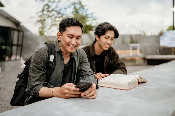 アジアの大学生2人が携帯電話で座って屋外のカフェで本を読む — ストック写真