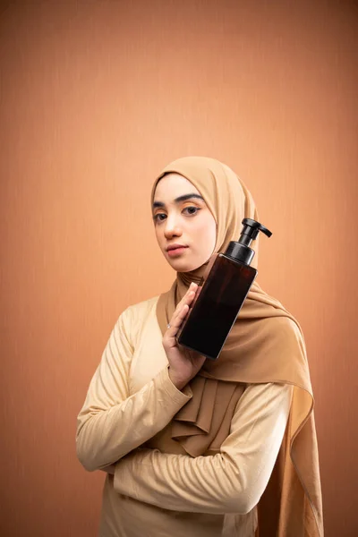 身穿头巾和奶油T恤的穆斯林妇女手牵着护肤瓶 在橙色的背景下对着相机微笑 — 图库照片