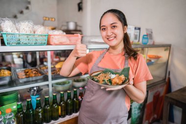 Güzel Asyalı kız, yemek standında bir tabak pekel yiyeceği taşıyarak başparmak kaldırıyor.