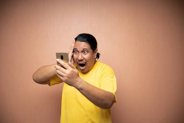 old asian man wearing yellow tshirt holding handphone and using it to texting or video call with each other in plain background isolated