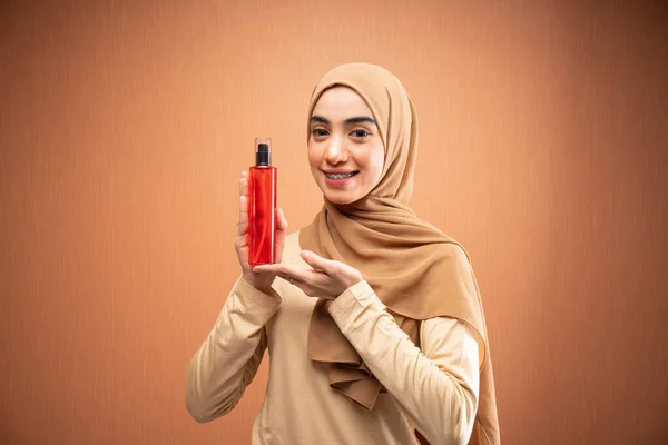 ヒジャーブとクリームTシャツを着たイスラム教徒の女性がオレンジの背景にカメラで微笑むスキンケアボトルを手に — ストック写真