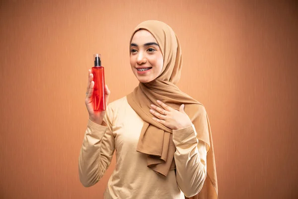 ヒジャーブとクリームTシャツを着たムスリム女性がオレンジ色の背景で横に笑顔でスキンケアボトルを抱えて胸に手を残しました — ストック写真