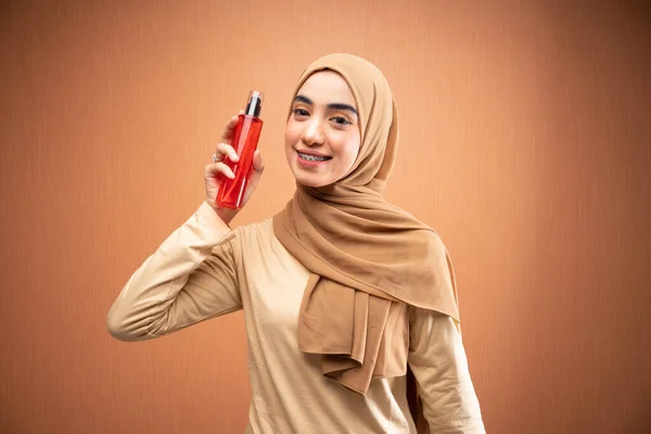 ヒジャーブとクリームTシャツを着たイスラム教徒の女性がオレンジの背景にカメラで微笑むスキンケアボトルを持っている — ストック写真