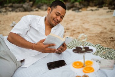 Genç adam piknik battaniyesinde oturmuş kitap okuyor ve tatilin tadını çıkarıyor.