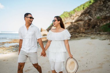 Beyaz elbiseli Asyalı çift sahilde el ele yürürken mutlu bir şekilde gülüyor.