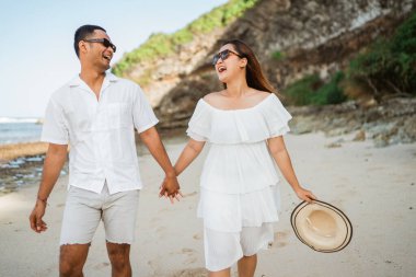 Beyaz elbiseli Asyalı çift kumsalda el ele yürürken