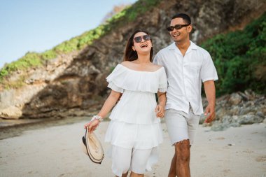 Beyazlı genç bir çift birlikte gülüyor ve tatilde sahilde yürüyor.