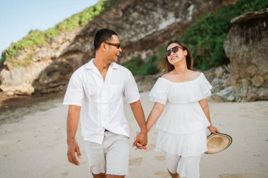 Beyaz elbiseli Asyalı çift kumsalda el ele yürürken