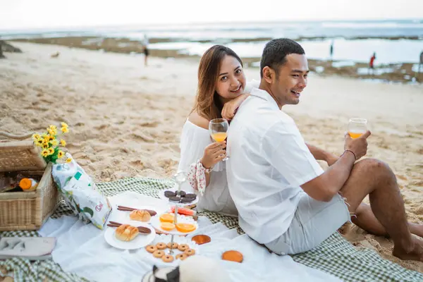 newlywed couple sitting on a picnic blanket toasting holding glasses during a picnic at the beach
