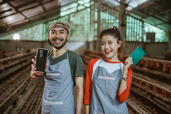 male and female workers show mobile phone screen and hold bankbooks standing at a chicken farm