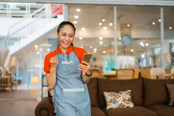 happy shop assistant woman in apron using mobile phone standing in front of furniture store