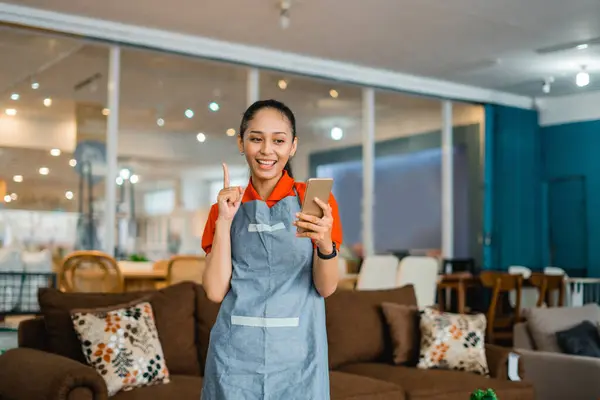 female shop assistant in apron using cell phone standing in front of furniture store