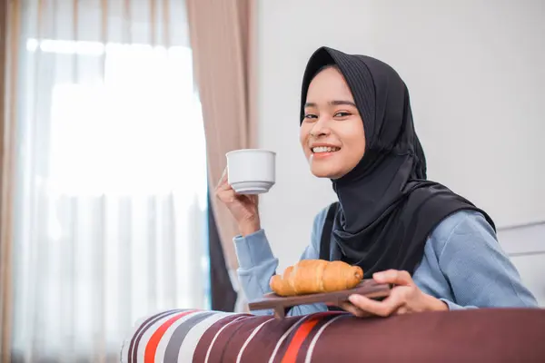 veiled woman smiling while having breakfast of bread and a cup of coffee sitting on the bed in the bedroom