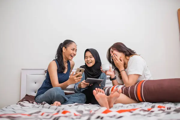 three women laugh at a cell phone and a digital tablet sitting on a bed