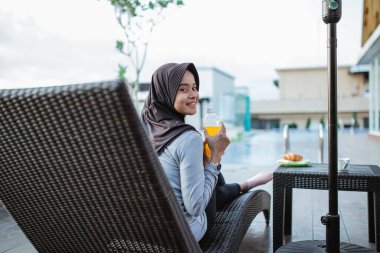 gülümseyen tesettürcü kız meyve suyu şişesini gösteriyor havuz kenarında oturuyor