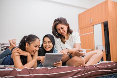 Üç kadın yatakta dijital tablet kullanırken gevşeyip sohbet ediyorlar.