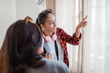 İki genç Asyalı kadın odanın perdeli penceresinden bir şeye bakıyor.
