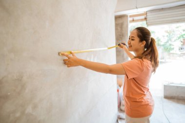 Asyalı genç bir kadın bir evin yapımına yardım ederken mezura kullanarak duvar ölçüsü ölçer.