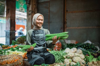 Önlüklü satıcı iki elinde de gülen yüzlü sebzeler tutuyor.