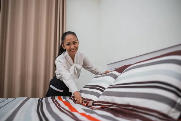 smiling hotel maid woman adjusting pillow on bed in hotel room