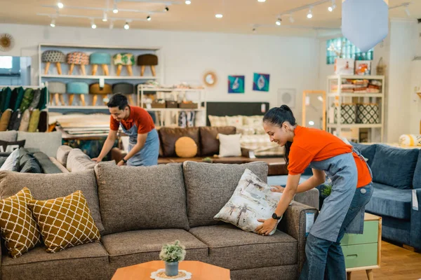 female shop assistant in apron tidying up sofa cushions during store opening at furniture store