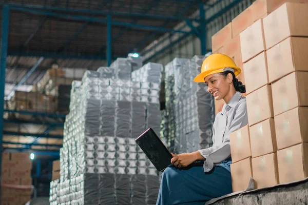 Asian woman factory worker wears safety helmet while sitting working with laptop in factory warehouse