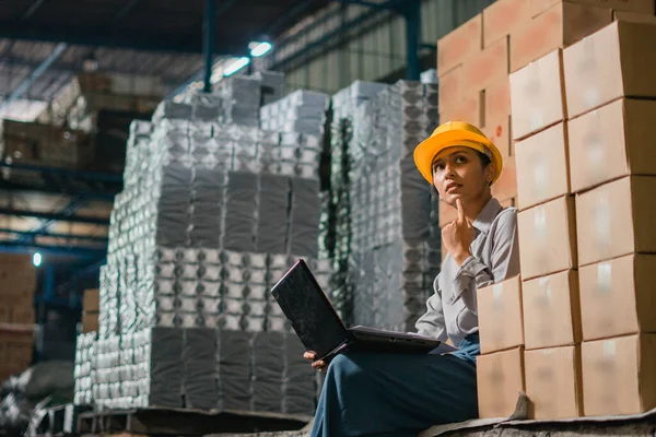 female factory worker sits near cardboard boxes wearing a safety helmet while working with a laptop computer in a factory