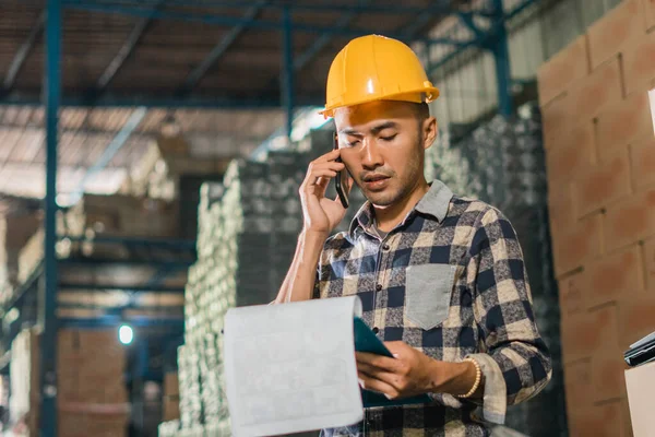 asian man factory worker on phone while working checking data of goods in storage warehouse