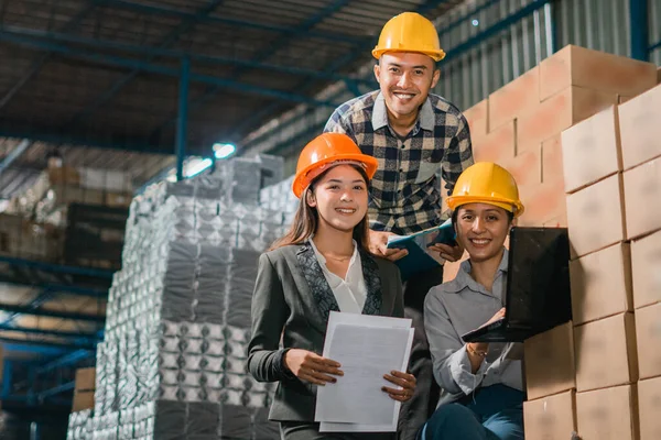 three factory employees smile at the camera while working on a laptop in the goods warehouse