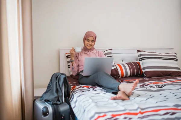 veiled woman drinking coffee with a cup and using a laptop on a bed in a hotel