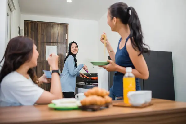 three Asian women laugh while making breakfast and eating together in the kitchen