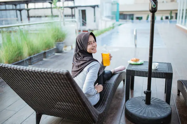 Gülümseyen Müslüman kadın havuz kenarında oturmuş meyve suyu şişesi gösteriyor.