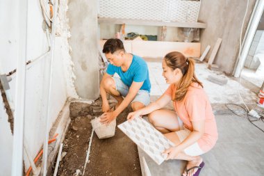 Asyalı genç kadın yeni bir ev inşa ederken kocanın fayans döşemesine yardım ediyor.