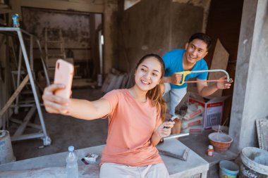 Karım, yeni bir ev inşa ederken kocasıyla sosyal medyada selfie çekmek için cep telefonu kullanıyor.