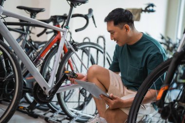 Bisiklet dükkanında dijital tablet taşıyan bisiklet pedallarının üzerine çömelmiş genç girişimci