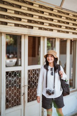 Bir turist yerine yürürken eski bir evin önünde kamera taşıyan sırt çantalı bir kadın turist duruyor.