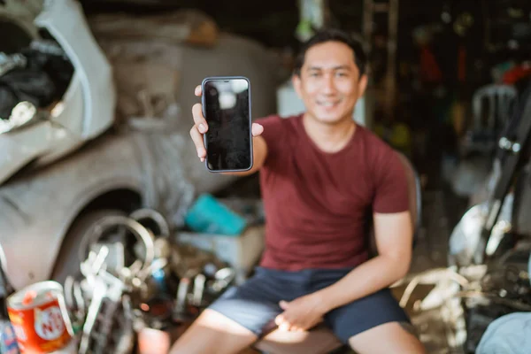 Handsome young mechanic shows a cell phone screen sitting on a chair in an old garage