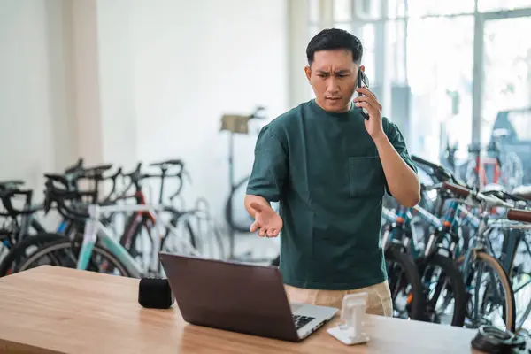 young man makes angry phone call while working on laptop at bike shop